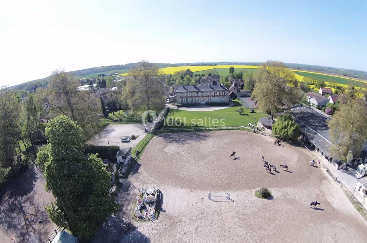 Vue aérienne d'un centre équestre avec une carrière, des cavaliers à cheval et des champs en arrière-plan.