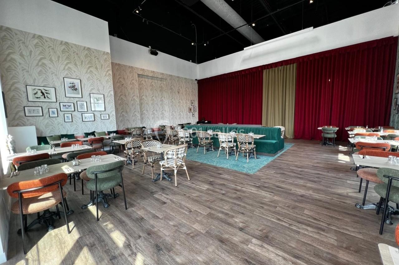 Salle de restaurant lumineuse avec parquet, tables dressées, chaises variées et rideaux rouges couvrant un mur.