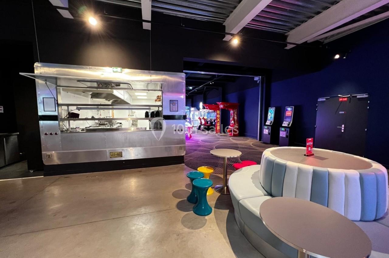 Espace intérieur avec coin salon moderne, tables colorées, cuisine ouverte et machines d'arcade en arrière-plan.