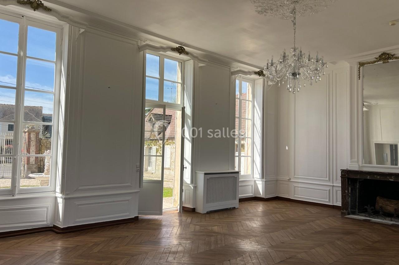 Salle lumineuse avec parquet en bois, grandes fenêtres, cheminée en marbre, miroir et lustre en cristal.