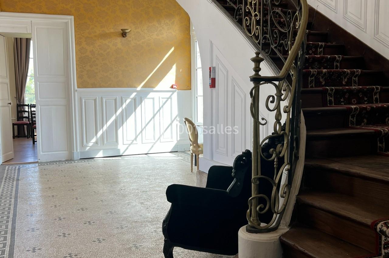 Escalier en bois avec rampe en fer forgé dans un hall lumineux, murs jaunes et blancs, sol en mosaïque.