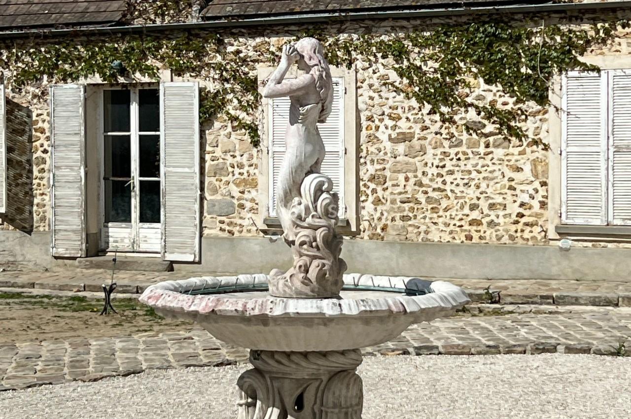 Statue en pierre représentant une femme, placée au centre d'une fontaine devant une maison en pierre avec volets blancs.