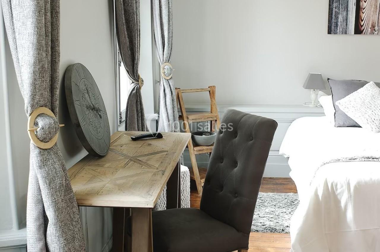 Bureau en bois avec chaise rembourrée dans une chambre lumineuse, décorée de rideaux gris et d'un lit blanc.
