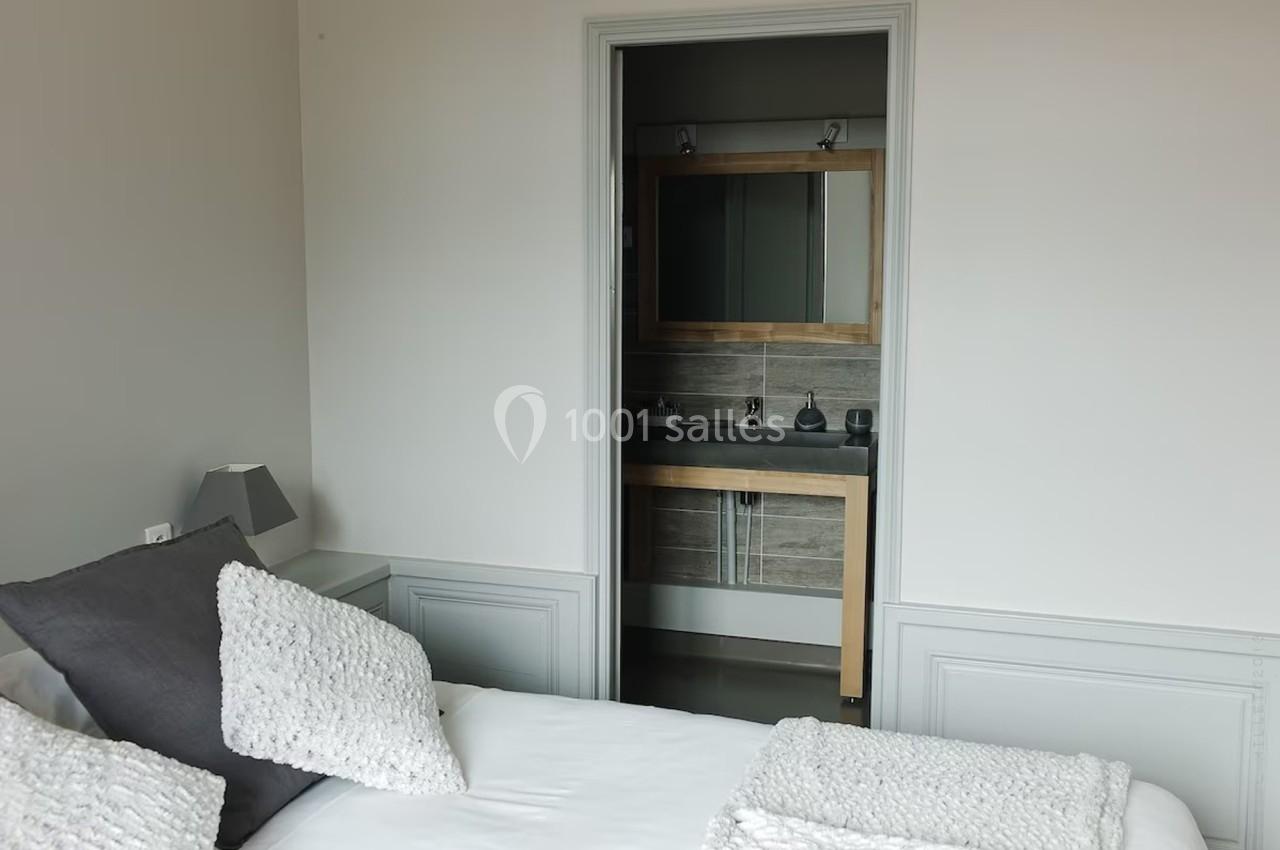 Chambre avec lit double, coussins blancs, et vue sur une salle de bain avec miroir et lavabo en arrière-plan.
