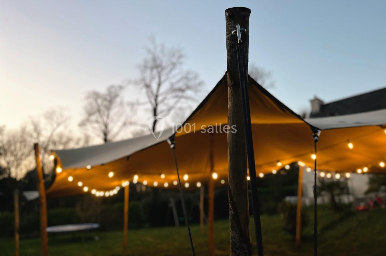Tente éclairée par des guirlandes lumineuses dans un jardin au crépuscule, avec des arbres en arrière-plan.