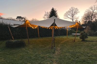 Tente beige éclairée par des guirlandes lumineuses, installée dans un jardin au coucher du soleil.