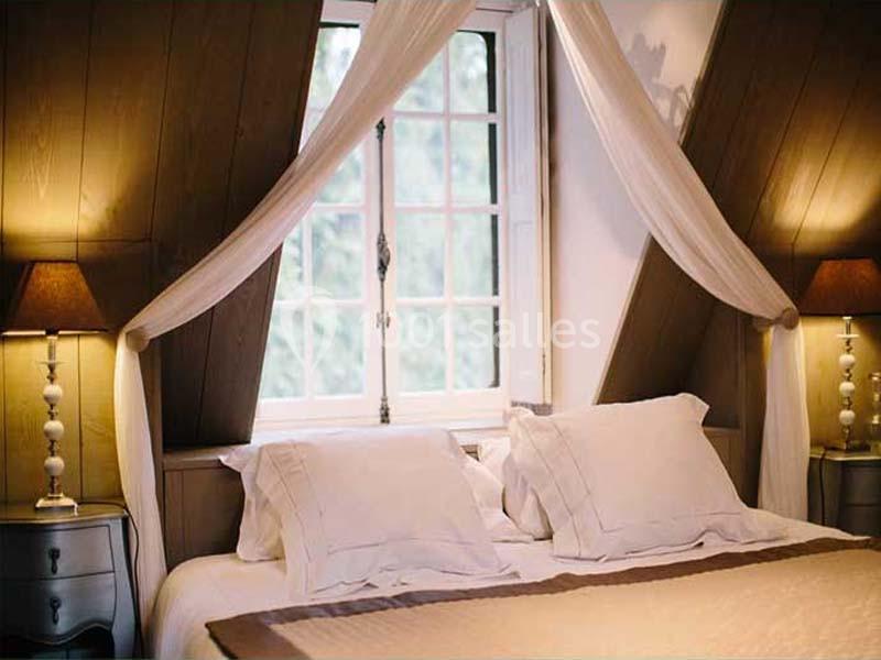 Lit double avec draps blancs, encadré par des rideaux légers, situé sous une fenêtre à croisillons dans une chambre…