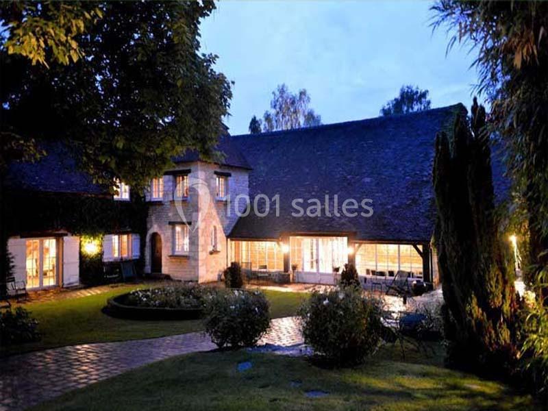 Maison en pierre éclairée à la tombée de la nuit, entourée d'un jardin avec allées pavées et arbres.