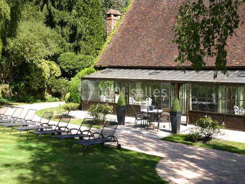 Jardin avec transats sur pelouse, chemin pavé et bâtiment avec verrière entouré de végétation.