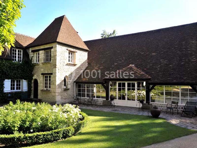 Façade d'une maison en pierre avec toit en pente, jardin fleuri et terrasse aménagée devant des baies vitrées.