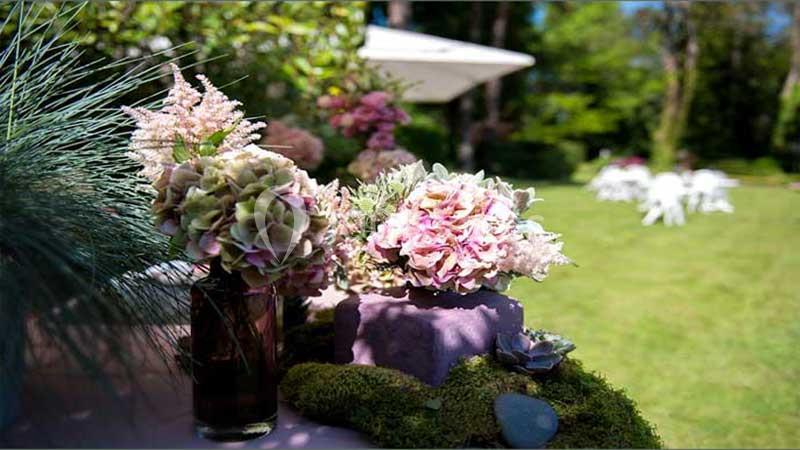 Arrangement floral avec hortensias roses et verdure sur une table, dans un jardin ensoleillé avec des chaises en arrière…
