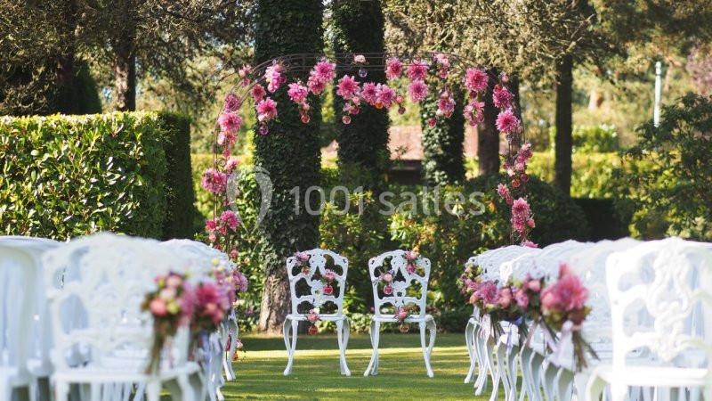 Chaises blanches décorées de fleurs roses alignées devant une arche florale dans un jardin verdoyant.