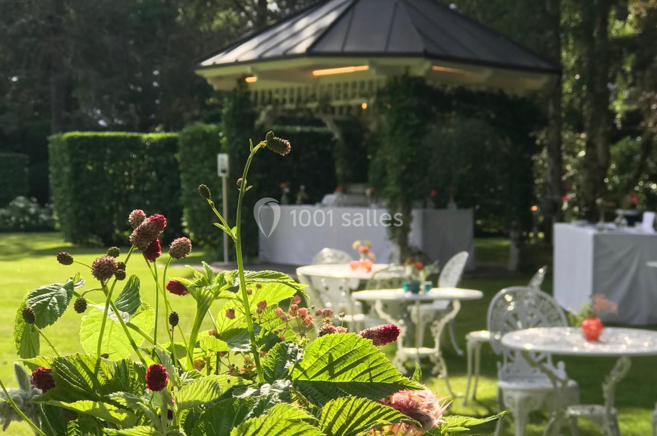Fleurs au premier plan avec un kiosque entouré de verdure et des tables blanches dans un jardin en arrière-plan.