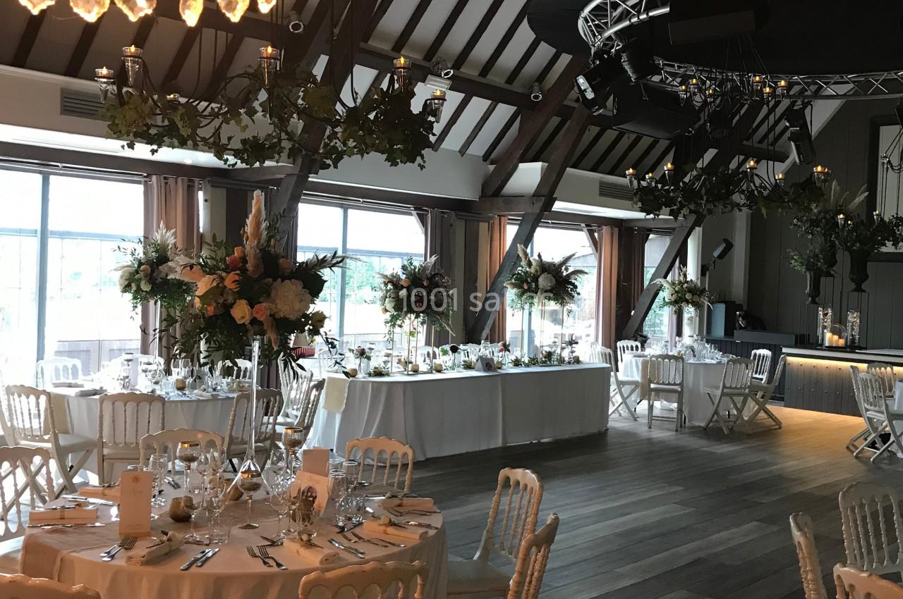 Salle de réception élégante avec tables décorées de fleurs, chaises blanches et grandes baies vitrées.