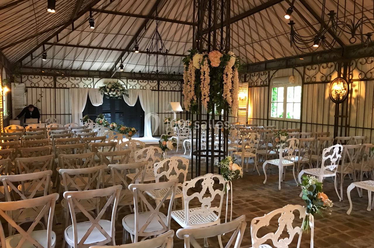 Salle décorée pour une cérémonie avec des chaises blanches alignées, des fleurs suspendues et un espace central dégagé.