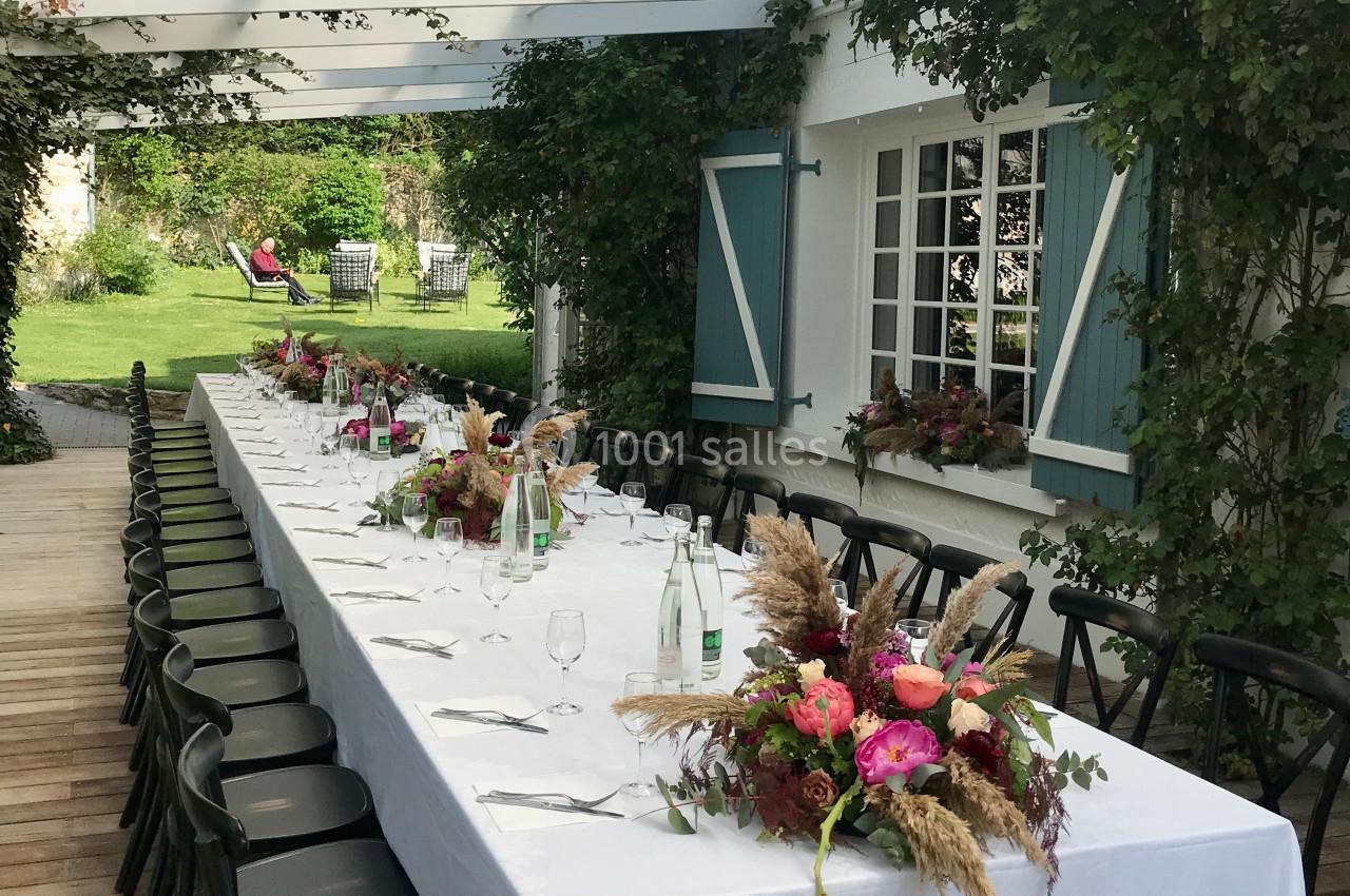 Grande table dressée en extérieur sous une pergola, ornée de fleurs et entourée de chaises noires, avec jardin en arrière…