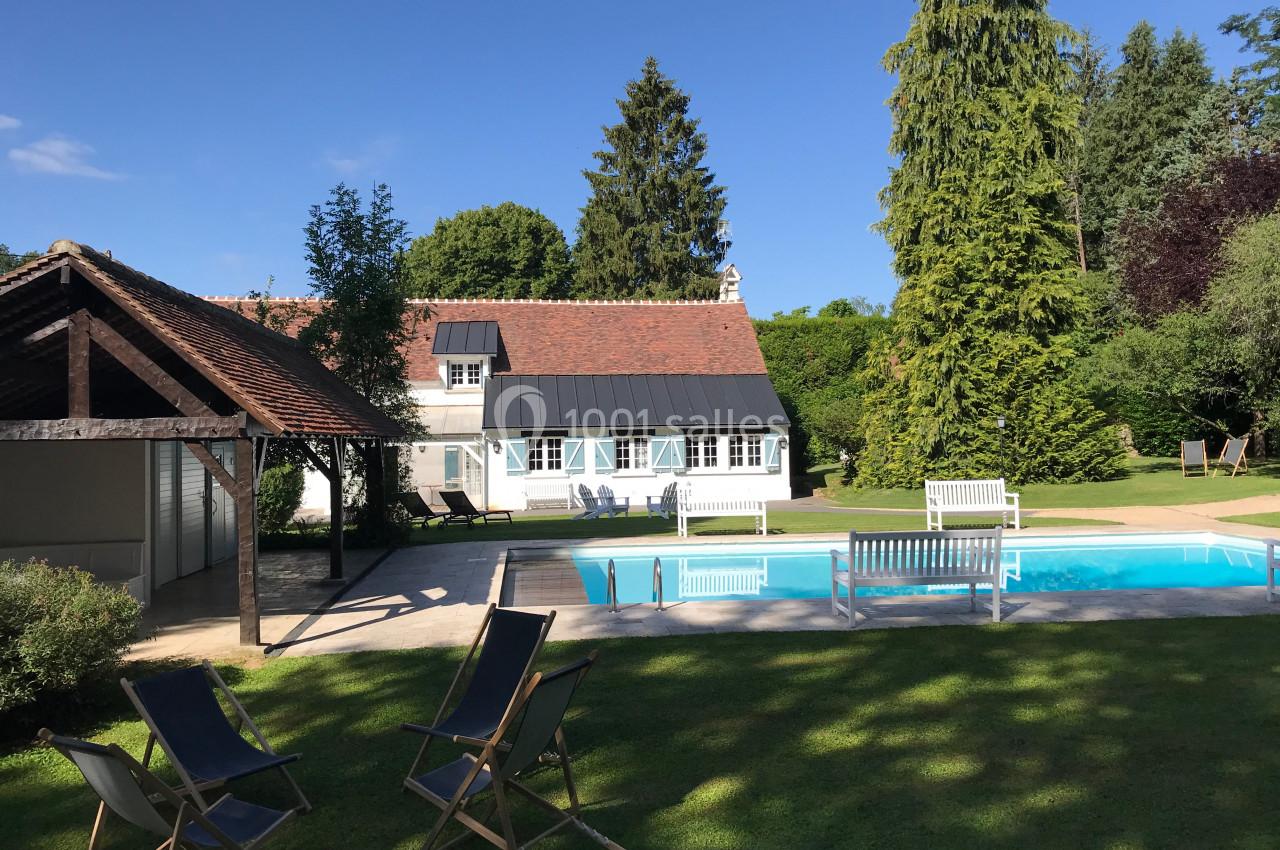 Maison avec piscine extérieure, transats sur une pelouse et jardin arboré sous un ciel bleu.