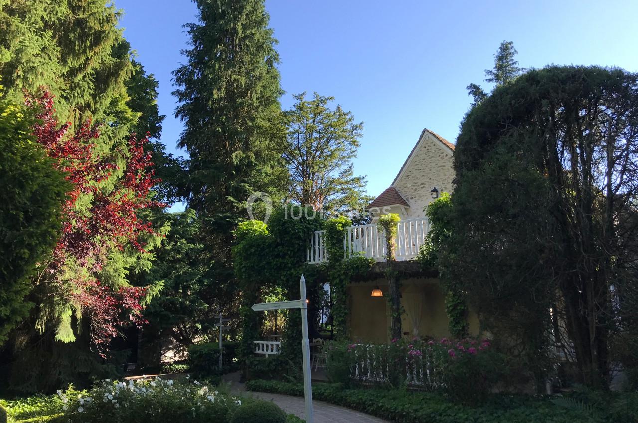 Jardin verdoyant avec allée pavée, maison en pierre entourée de végétation et ciel bleu dégagé en arrière-plan.