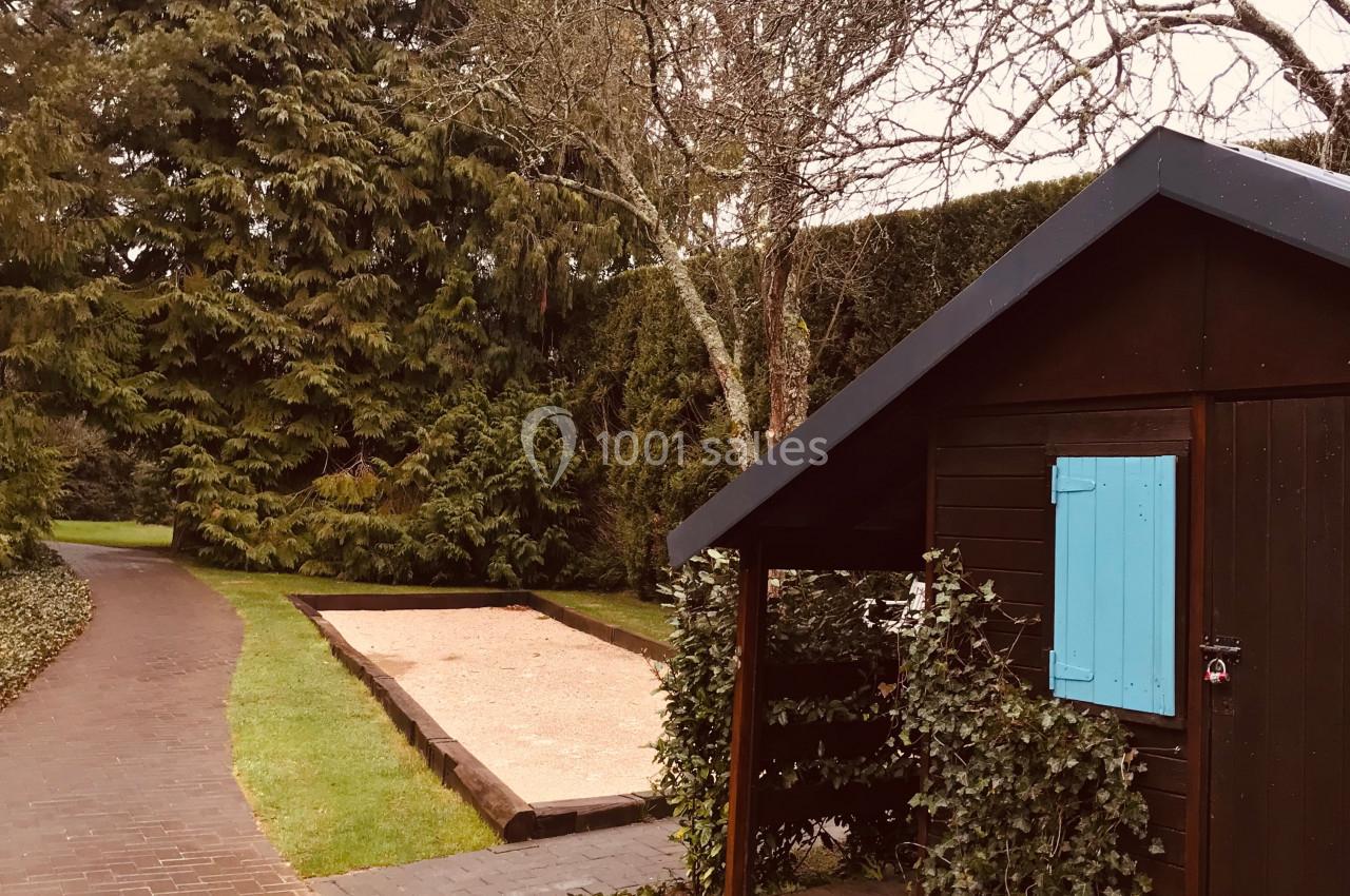 Abri en bois avec volet bleu, entouré de végétation, près d'une allée pavée et d'un terrain de pétanque.
