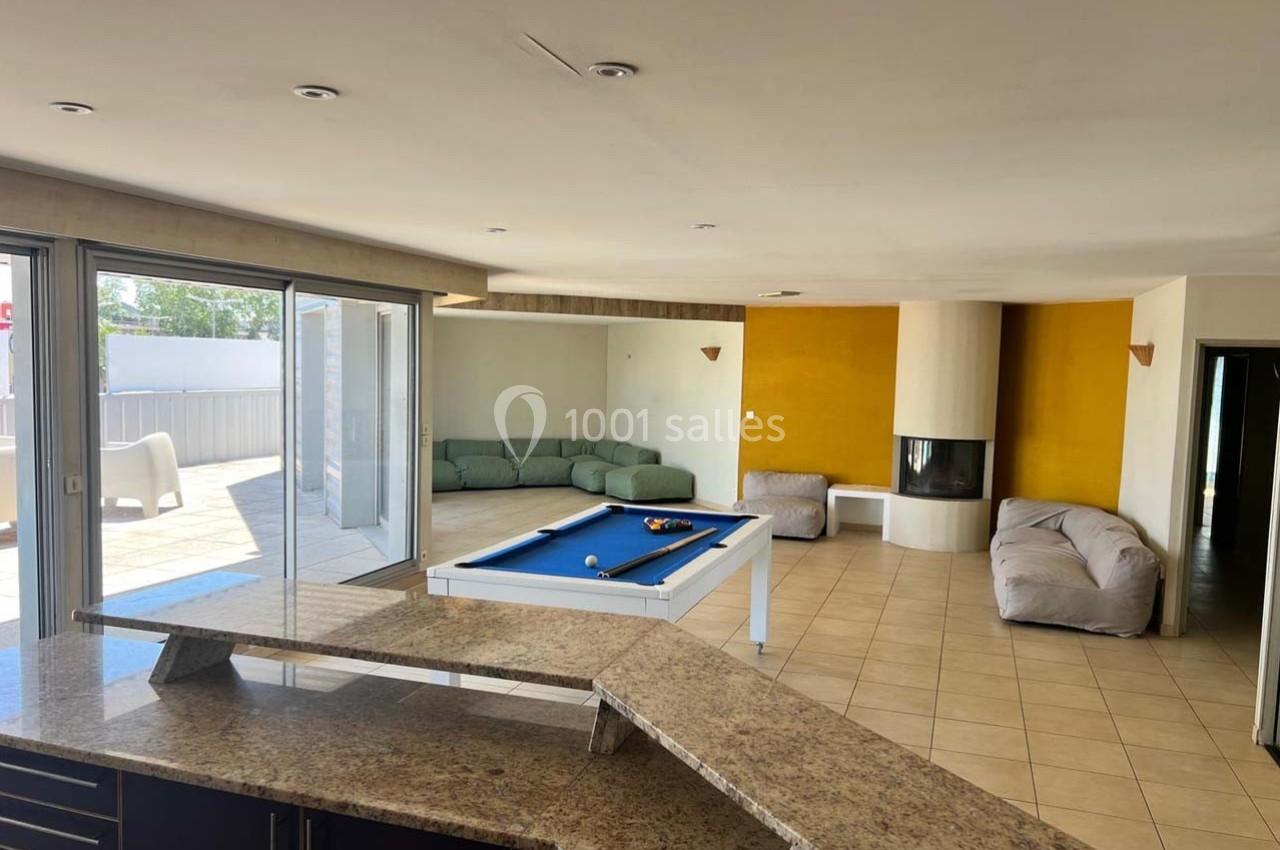 Salle lumineuse avec billard, canapé d'angle, cheminée moderne, et grande baie vitrée donnant sur une terrasse.