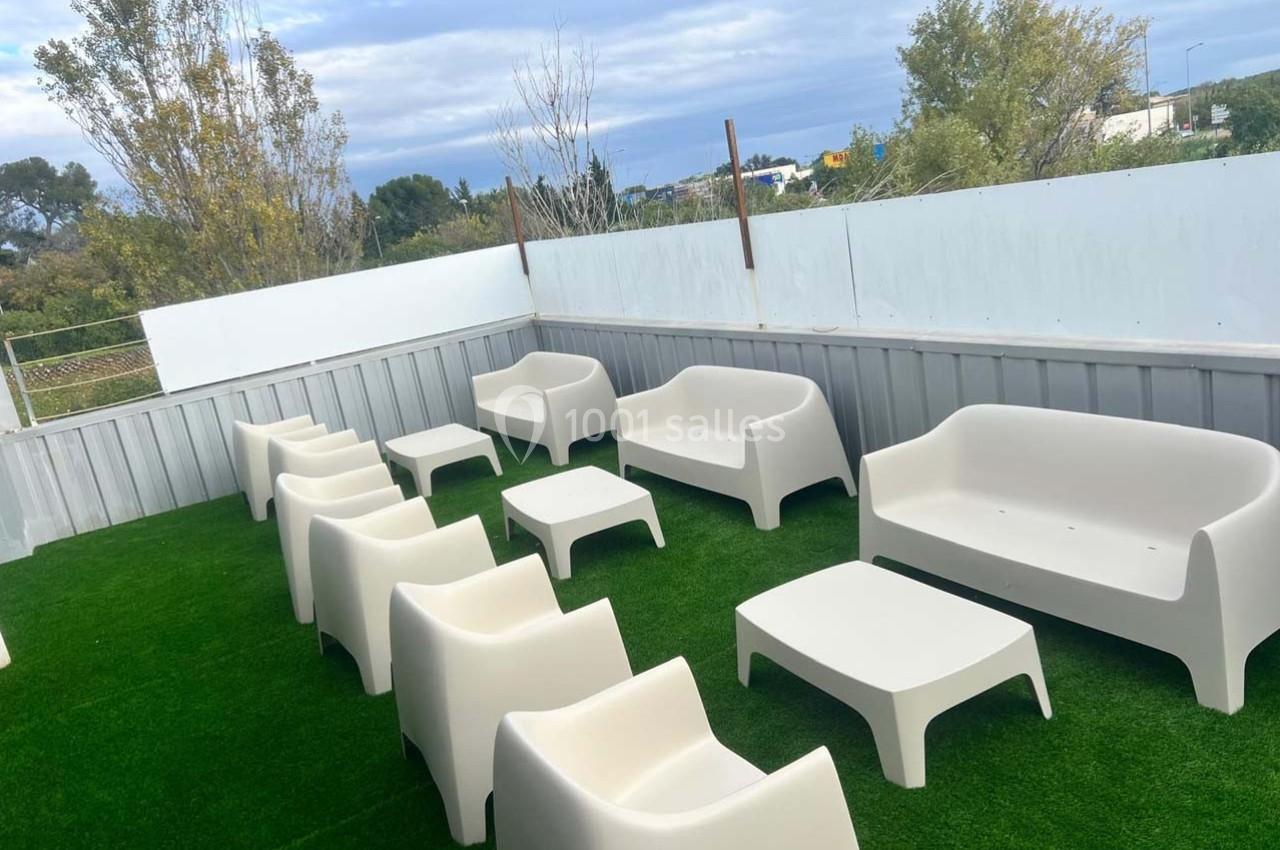 Mobilier de jardin blanc disposé sur une terrasse avec pelouse artificielle, entourée de panneaux et d'arbres en arrière…