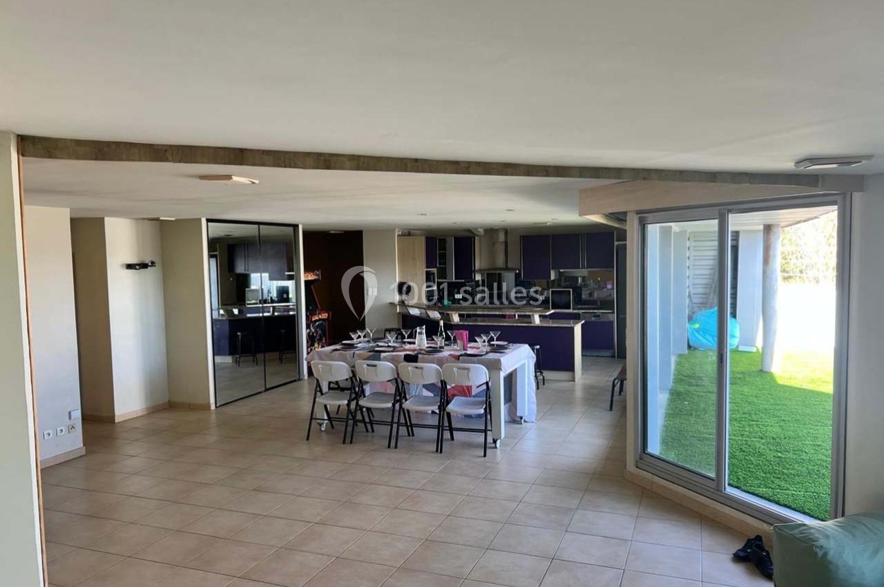 Salle à manger ouverte avec table dressée, cuisine moderne en arrière-plan et grande baie vitrée donnant sur un jardin.