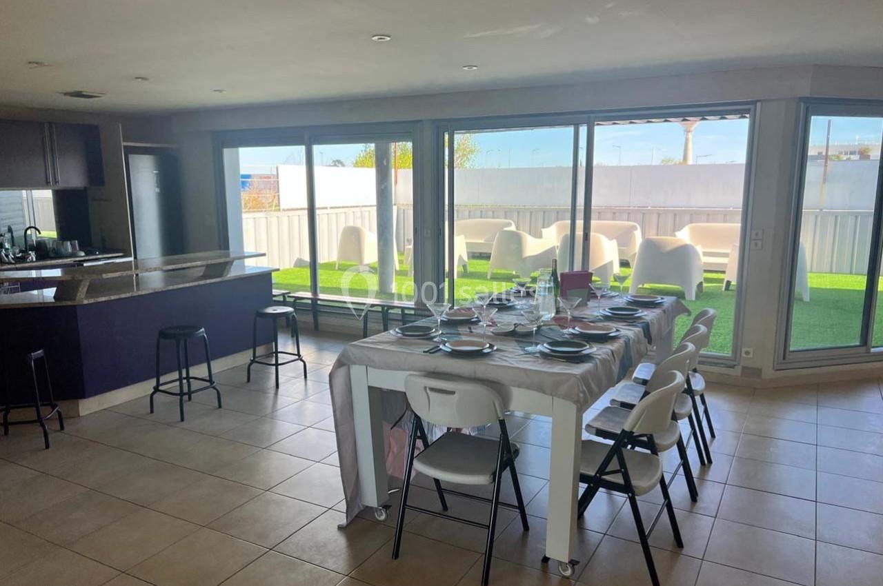 Salle à manger lumineuse avec une grande table dressée, cuisine ouverte et accès à une terrasse extérieure.