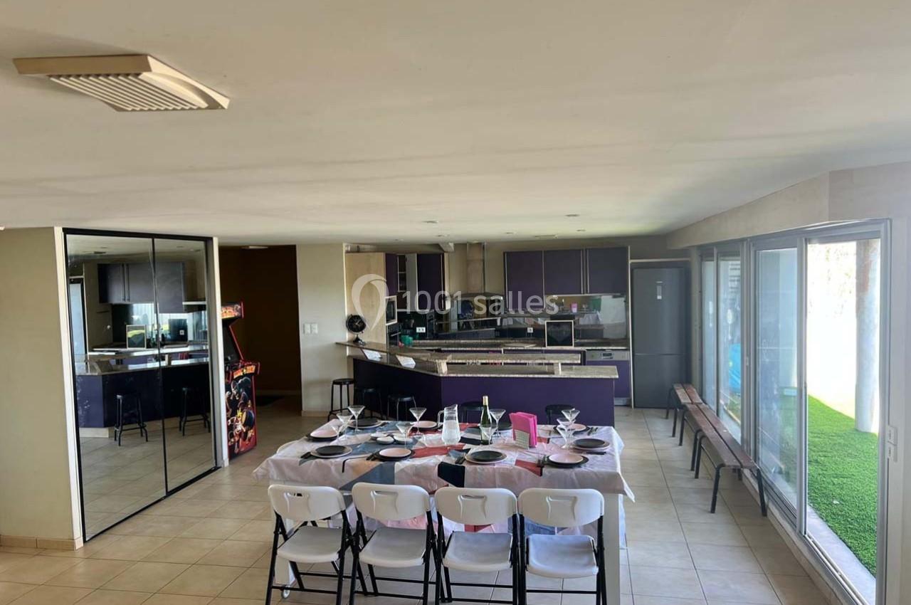 Salle à manger avec table dressée, chaises pliantes blanches, cuisine ouverte moderne et grandes baies vitrées.