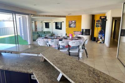 Vue d'un grand salon lumineux avec carrelage beige, cuisine ouverte et baies vitrées donnant sur un jardin.