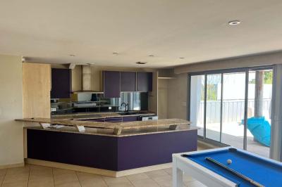 Vue d'un grand salon lumineux avec carrelage beige, cuisine ouverte et baies vitrées donnant sur un jardin.