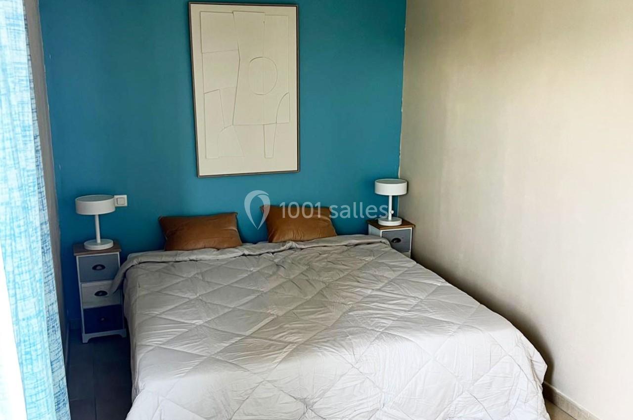 Lit double avec couette blanche, deux tables de chevet avec lampes et mur bleu décoré d'un tableau abstrait.