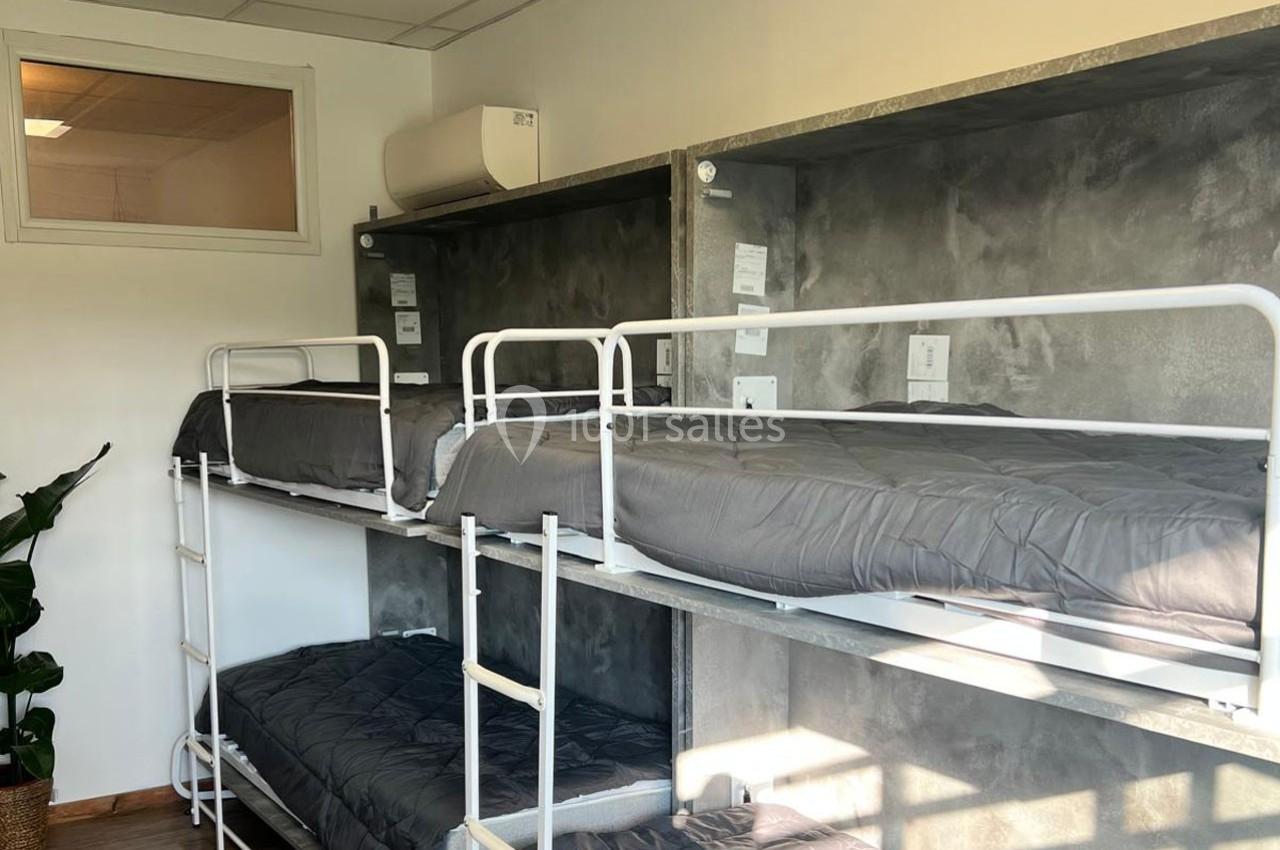 Dortoir avec lits superposés en métal, draps gris et mur en béton, éclairé par une fenêtre intérieure.