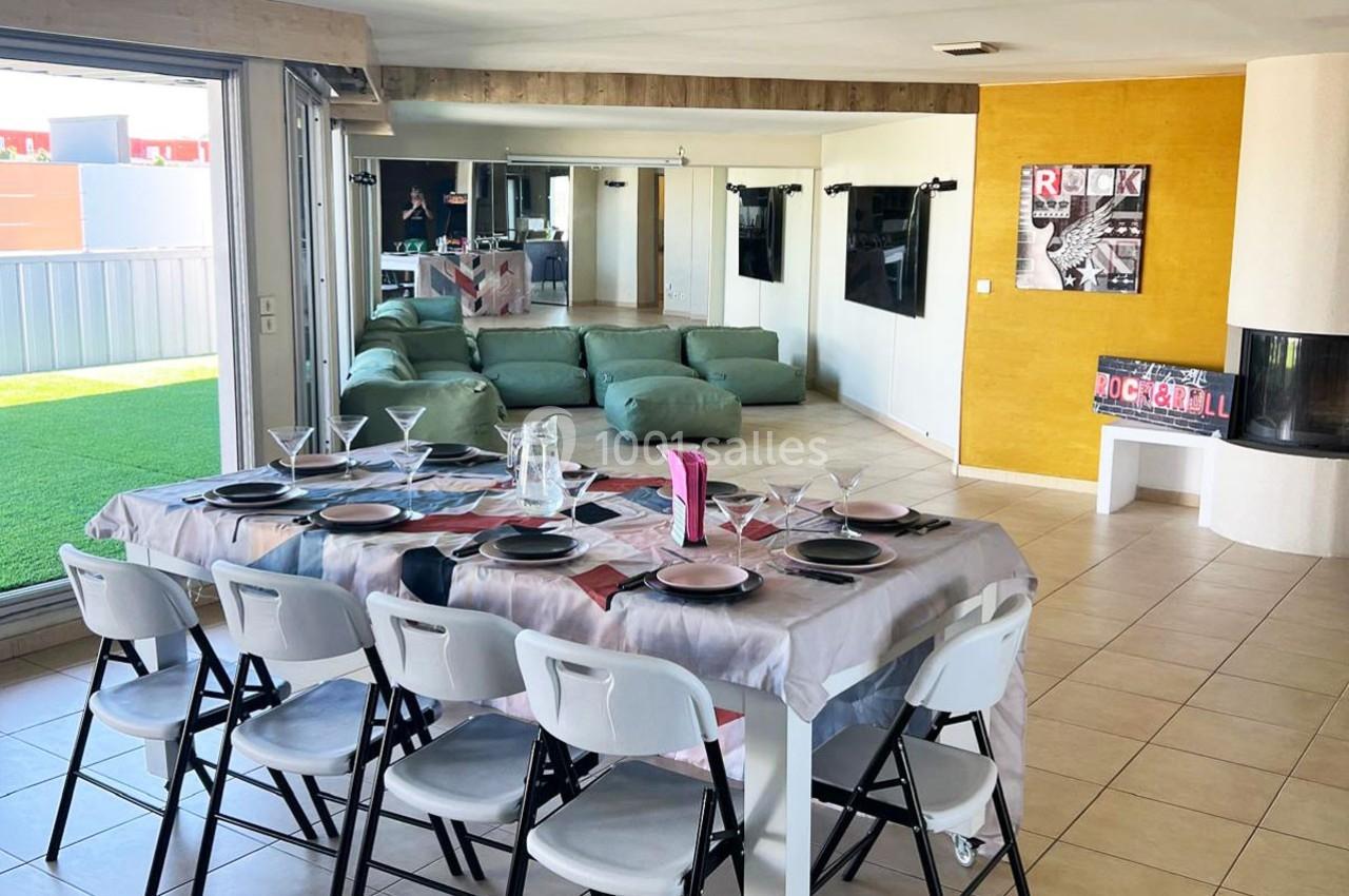Salle à manger moderne avec table dressée, chaises pliantes blanches, canapé modulable et accès à une terrasse extérieure.