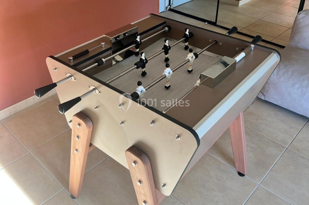 Table de baby-foot beige avec joueurs noirs et blancs, posée sur un sol carrelé près d'un mur rouge.