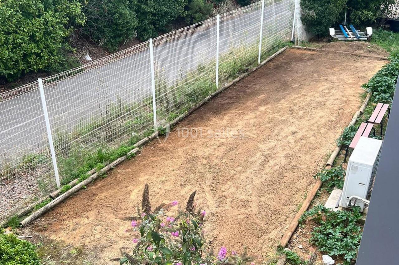 Terrain en terre battue entouré de végétation, bordé par une clôture grillagée et une route en arrière-plan.