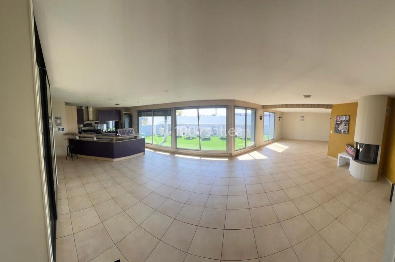 Vue d'un grand salon lumineux avec carrelage beige, cuisine ouverte et baies vitrées donnant sur un jardin.