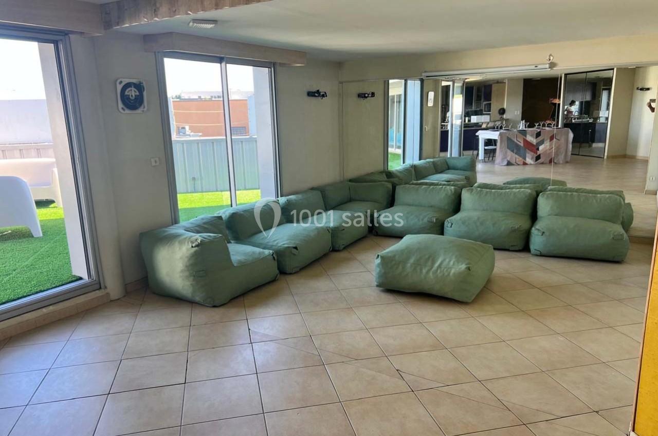 Des poufs verts disposés en forme de U dans une pièce lumineuse avec vue sur une terrasse et une cuisine ouverte.