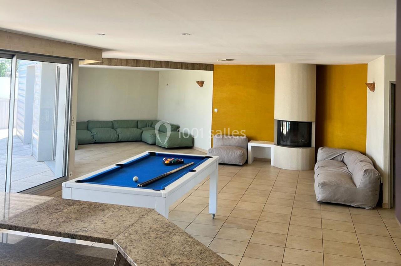 Salle lumineuse avec table de billard, canapés, cheminée moderne et grandes baies vitrées donnant sur une terrasse.