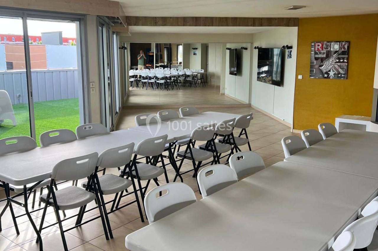 Salle lumineuse avec de grandes tables et des chaises pliantes blanches, donnant sur un espace extérieur avec pelouse.