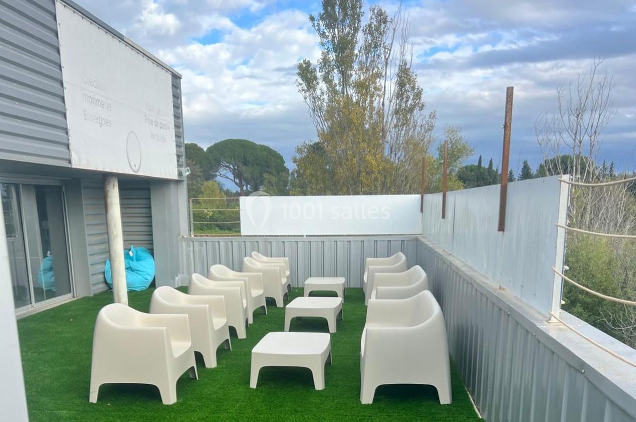 Terrasse extérieure avec fauteuils et tables blanches sur gazon synthétique, entourée de barrières métalliques.