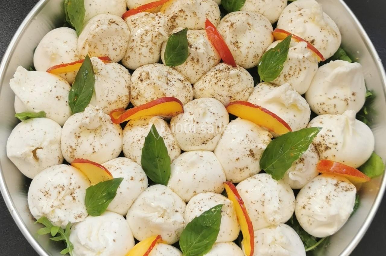 Assortiment de boules de mozzarella garnies de feuilles de basilic, tranches de pêche et épices, sur un lit de verdure.