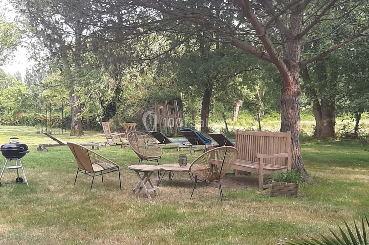 Mobilier de jardin en bois et métal disposé sur une pelouse ombragée, avec un barbecue et des transats en arrière-plan.