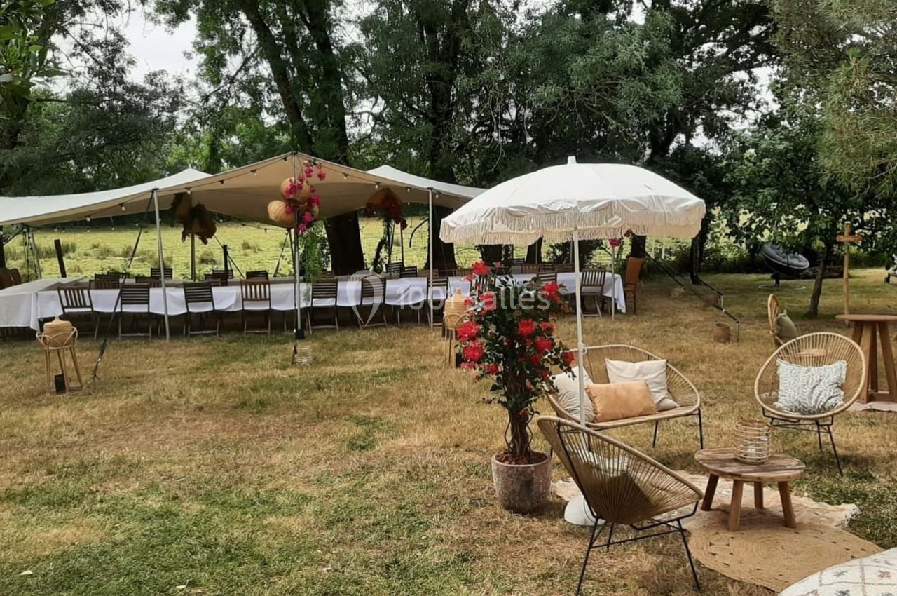 Tables dressées sous une tente et un parasol dans un jardin, entourés de chaises, de fleurs et de mobilier en rotin.