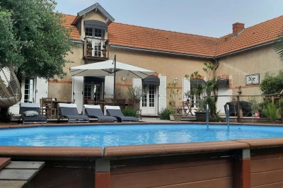 Maison en pierre entourée de verdure, avec un chemin de gravier, des arbres et un jardin en premier plan.