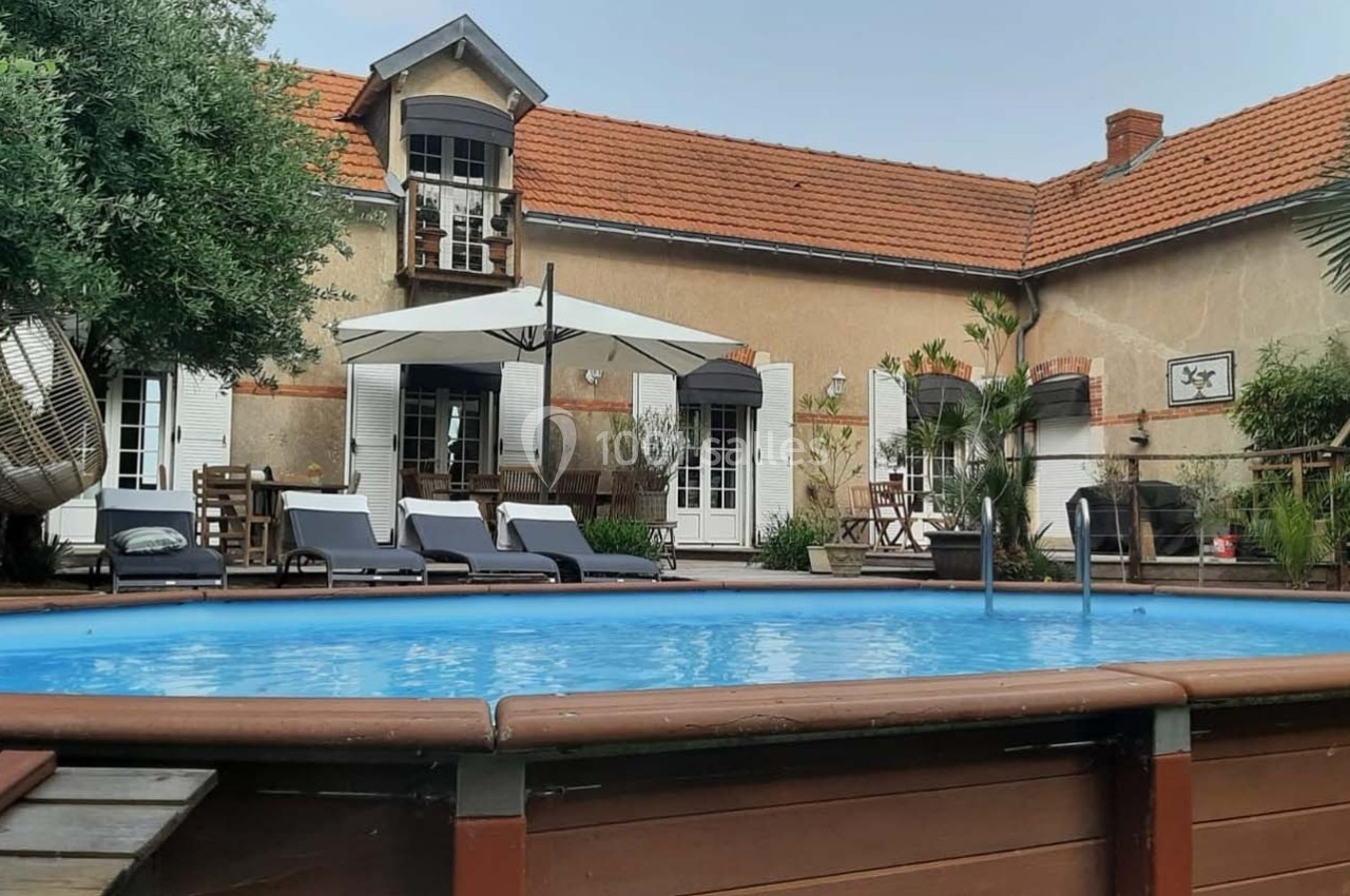 Maison avec terrasse, piscine hors-sol entourée de bois, chaises longues et parasol dans un jardin arboré.