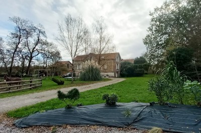 Maison en pierre entourée de verdure, avec un chemin de gravier, des arbres et un jardin en premier plan.