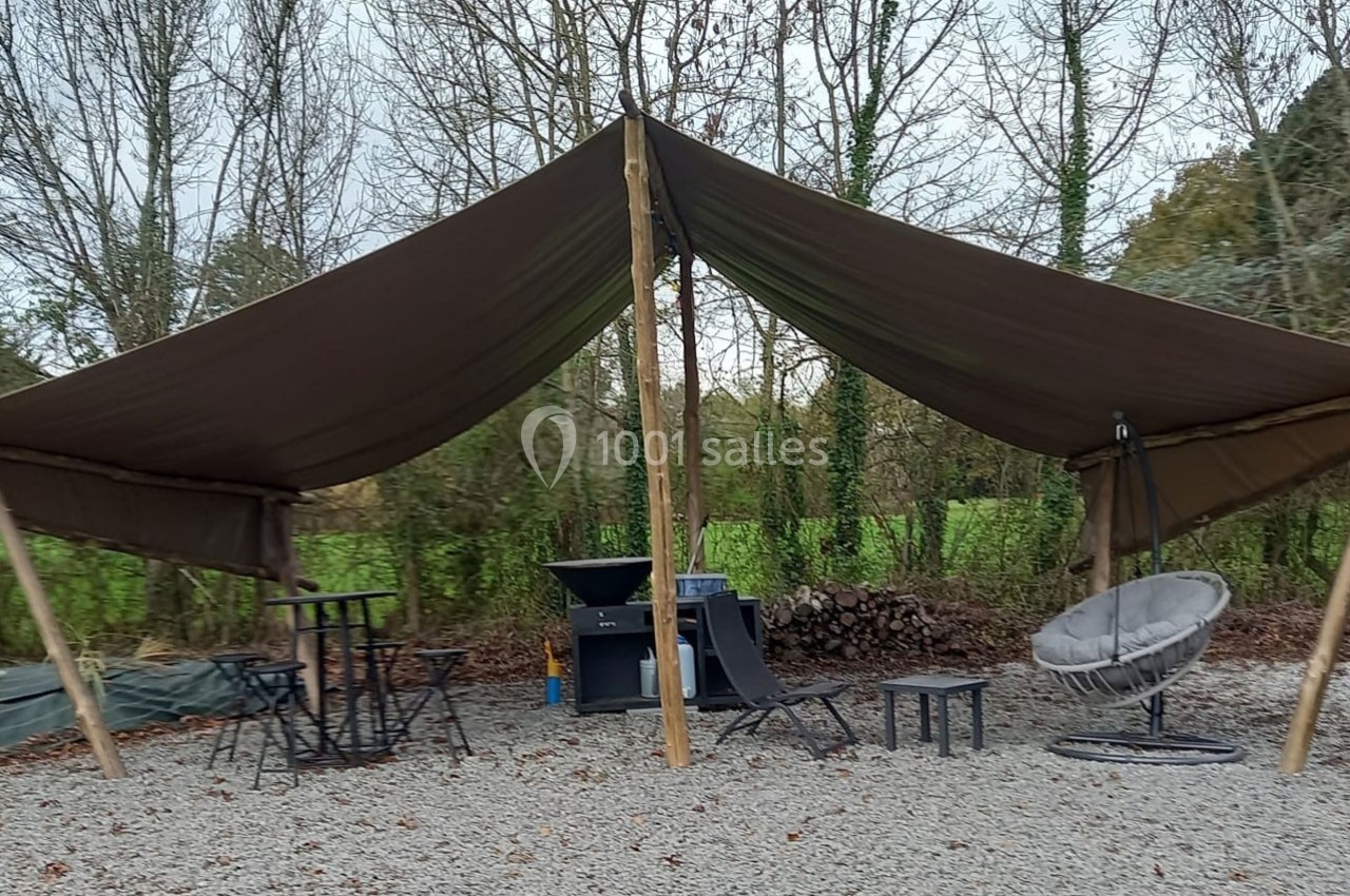 Tente en toile marron montée sur des poteaux, abritant des meubles de jardin sur un sol en gravier, entourée d'arbres.