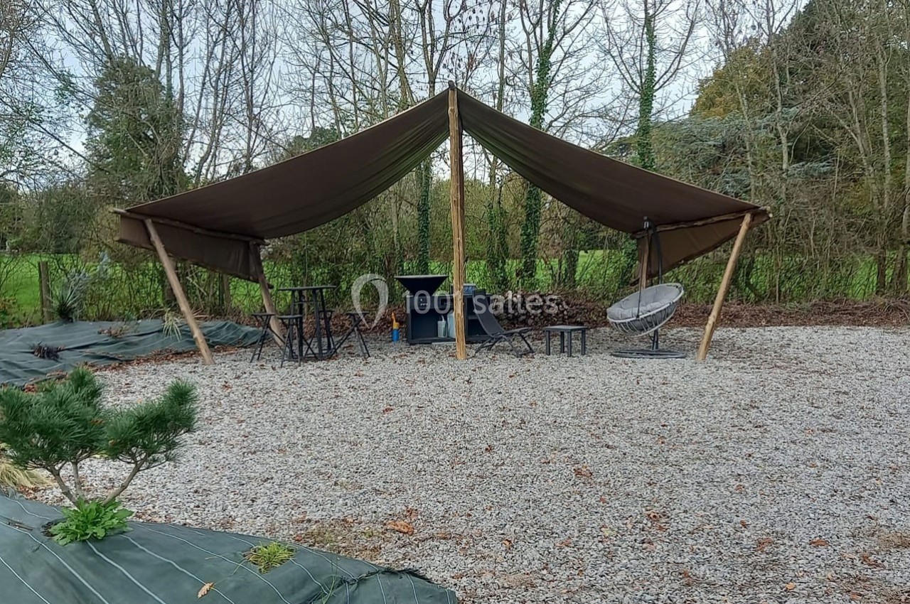 Abri en toile marron sur une zone gravillonnée, avec mobilier de jardin et bois empilé en arrière-plan.