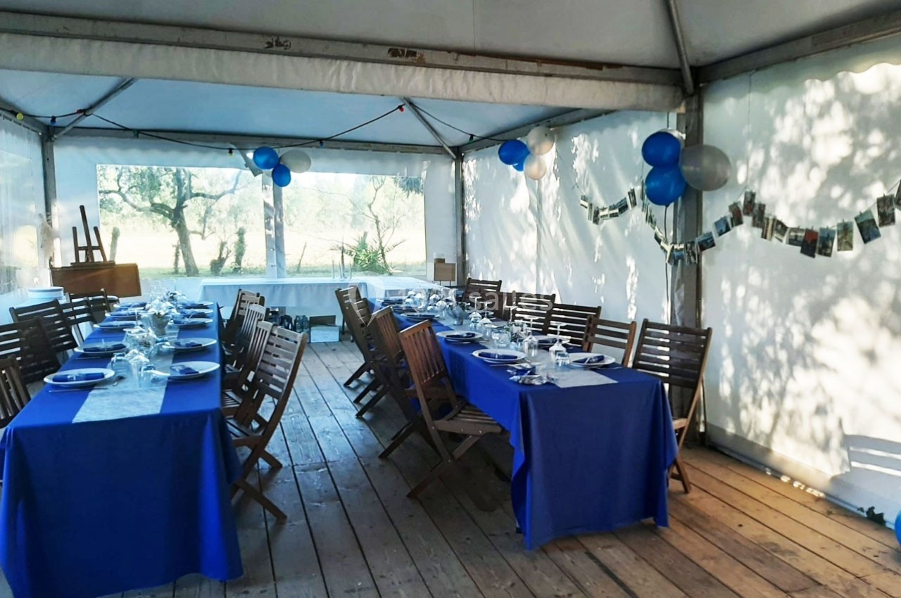 Tables dressées avec nappes bleues et chaises en bois dans une tente lumineuse décorée de ballons et guirlandes de photos.