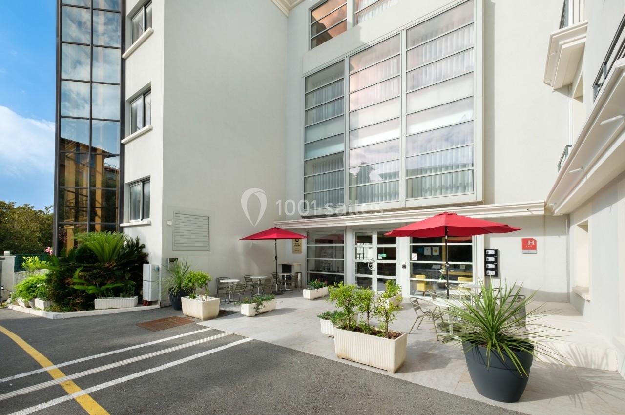 Entrée d'un bâtiment moderne avec terrasse aménagée, tables, chaises et parasols rouges, entourée de plantes en pots.